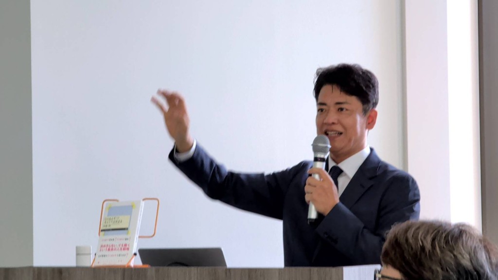 演台の前でマイクを持ち、手を挙げながら報告する中嶌剛